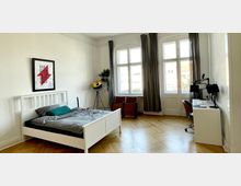 Ein helles Schlafzimmer mit großem Fenster und grauen Vorhängen, ausgestattet mit einem weißen Bett, einem Schreibtisch mit Monitoren und einer Stehlampe. Der Raum hat Parkettboden, helle Wände und dekorative Elemente wie ein gerahmtes Poster an der Wand.