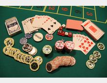 Spielszene auf einem grünen Casinotisch mit Karten, Pokerchips verschiedener Werte, roten Würfeln und zwei Kartenstapeln in Verpackungen. Ein Dealer-Chip liegt zentral zwischen den Spielutensilien.