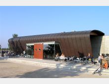 Modernes Cafégebäude mit geschwungener Metallfassade in dunklem Braun und einer Glasfront, davor eine Terrasse mit schwarzen Stühlen und Tischen, an denen Menschen sitzen; umgeben von einer offenen, parkähnlichen Umgebung bei klarem Himmel.