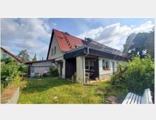 Einfamilienhaus mit rotem Satteldach, großem Balkon und hellen Fassaden, umgeben von einem Garten mit Wiese und grünen Sträuchern. Im Vordergrund ist eine offene Terrasse mit Gartenmöbeln zu sehen.