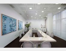 Konferenzraum mit U-förmig angeordneten Tischen, weißen Wänden und einer abgehängten Decke mit eingebauten Leuchten. An der Wand hängen zwei abstrakte Kunstwerke, im Raum stehen graue Stühle, eine große Topfpflanze, sowie eine weiße Kommode mit Orchideen im Hintergrund.