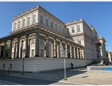 Neoklassizistisches Gebäude mit Säulengang und weißen Fassaden, durchzogen von verzierenden Details. Im Vordergrund ist eine gepflasterte Straße und ein Schild mit einer Geschwindigkeitsbegrenzung von 30 km/h sichtbar.