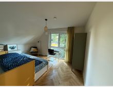 Ein schlicht eingerichtetes Schlafzimmer mit einem gelben Bettgestell, Parkettboden und einer Dachschräge. Ein Fenster mit gelben Vorhängen bietet Blick ins Grüne; daneben stehen ein Schreibtisch mit Stuhl sowie ein Kleiderschrank.