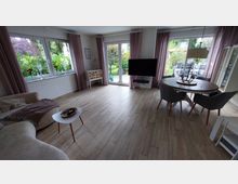 Helles Wohnzimmer mit großen Fenstern und Terrassentüren, die einen Blick in den Garten bieten. Der Raum ist mit einem beigen Sofa, einem runden Esstisch mit grauen Stühlen und einem Wand-TV ausgestattet; der Boden ist mit hellen Holzfliesen gestaltet.