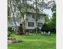 Zweistöckiges Haus mit weißer Fassade und braunen Klinkerelementen, umgeben von einem grünen Garten mit Rasenfläche, Bäumen und Gartenmöbeln. Im Vordergrund stehen Tische und Stühle, sowie einzelne Blumenbeete.
