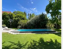 Ein Garten mit Rasenfläche, einem rechteckigen Swimmingpool mit angrenzendem Holzdeck, einem Basketballkorb und umliegenden Bäumen und Büschen. Im Hintergrund ist ein Haus mit einem roten Ziegeldach sichtbar.