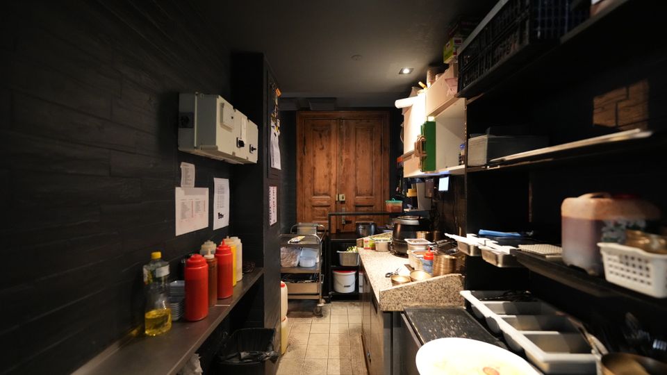 Schmaler Küchen- und Vorbereitungsraum mit dunklen Wänden, Fliesenboden und Arbeitsfläche aus Stein, flankiert von Regalen mit Behältern, Küchenutensilien und Gewürzflaschen. Am Ende des Raums steht ein großer Holzschrank unter Deckenbeleuchtung.