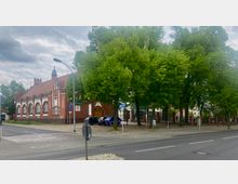 Backsteingebäude mit gotischen Elementen und zahlreichen Rundbogenfenstern, flankiert von hohen Bäumen entlang der Straße; im Vordergrund Parkplätze und Verkehrszeichen, im Hintergrund ein umzäuntes Gelände.