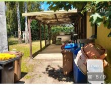 Überdachte Terrasse mit Holzpfeilern, daneben mehrere Mülltonnen und Abstellgegenstände. Im Hintergrund ein Garten mit Bäumen und Sträuchern.