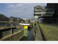 Industriehafen mit alten Lagerhäusern aus Backstein entlang eines Kanals. Im Vordergrund liegen Lastkähne im Wasser, daneben eine gepflasterte Uferpromenade mit Grasbewuchs, darüber ragt ein alter Kran mit Gerüst hervor.