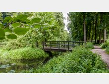 Holzbrücke über einen kleinen Bach in einer grünen Parklandschaft mit dichtem Laub, umgeben von hohen Bäumen und einem schmalen Fußweg rechts vom Wasserlauf.