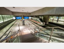 Unterirdischer Bahnhof mit modernen Zügen und heller Beleuchtung. Ein ICE fährt auf einem der Bahnsteige ein, daneben große Uhren, Anzeigetafeln und eine Treppe mit gläsernem Geländer im Vordergrund.