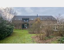 Steinbauernhaus mit Satteldach, großer zentraler Glasbalkon und mehreren Fenstern, umgeben von einem Garten mit Rasen, Büschen und kahlen Bäumen unter bewölktem Himmel. Rechts befindet sich ein Anbau aus Glas.