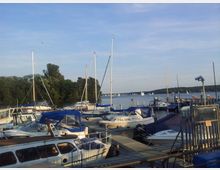 Jachthafen mit mehreren Segel- und Motorbooten, die an Stegen festgemacht sind, im Vordergrund. Im Hintergrund ein See mit weiteren Segelbooten sowie bewaldete Ufer. Der Himmel ist klar und lichtblau.