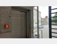 Ein moderner Innenraum mit einem Aufzug aus Edelstahl in einer Betonwand; daneben ein orangefarbener Notrufschalter. Durch große Fensterfronten ist ein Blick auf einen gepflasterten Hof mit parkenden Autos und umliegenden Gebäuden sichtbar.