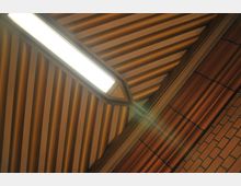 Decke mit diagonalen, goldfarbenen Metallrippen, einem geometrischen Ausschnitt in der Mitte und einer rechteckigen, leuchtenden Lampe. Rechts sind Ziegelwände mit vertikalen und horizontalen Linien zu sehen.