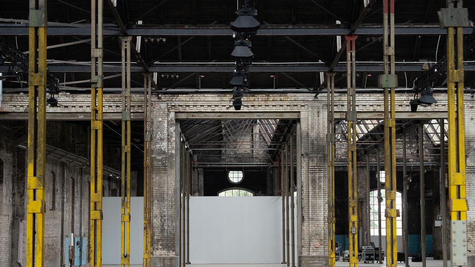 Interior of a large empty industrial warehouse with exposed steel roof trusses, hanging light fixtures, and rows of tall yellow support columns. Weathered brick walls and a wide open concrete floor lead toward a white partition wall at the far end.