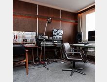Aufnahme eines Tonstudios mit holzvertäfelter Wand, Misch- und Synthesizer-Modulen, Bandmaschine sowie Mikrofonständer. Rechts stehen ein Schreibtisch mit Monitor und Lautsprecher neben einem großen Fenster mit Vorhängen; auf dem Boden liegen zahlreiche Kabel.