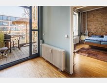 Schlafzimmer mit Holzboden und sichtbarer Backsteinwand, daneben ein offener Bereich mit Zugang zu einem Balkon mit Holzdielen, Grill und einem Sonnenschirm aus Stroh. Ein großer Heizkörper befindet sich an der Wand zwischen den Räumen.