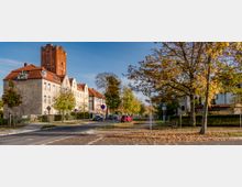 Straßenszene in einem Wohngebiet mit herbstlicher Stimmung; links stehen mehrstöckige Gebäude mit roten Ziegeldächern, im Hintergrund ein hoher, rotbrauner Backsteinturm. Rechts säumen Bäume mit verfärbten Blättern den Gehweg, einige parkende Autos und eine Sitzbank sind ebenfalls sichtbar.