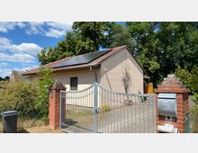 Ein einstöckiges Wohnhaus mit hellbeiger Fassade und bronzefarbenem Ziegeldach, auf dem Solarpaneele installiert sind, umgeben von Bäumen und Sträuchern. Im Vordergrund befindet sich ein Metalltor mit Backsteinpfosten, eines davon hat einen Briefkasten.