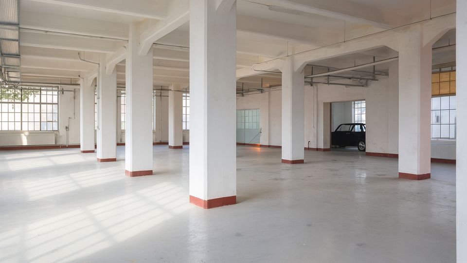 Großer, leerer Industrie- oder Lagerraum mit glattem Betonboden, vielen weißen Stützsäulen und sichtbaren Deckenleitungen. Hohe, rasterförmige Fenster lassen viel Tageslicht herein; im Hintergrund steht ein einzelnes Auto.