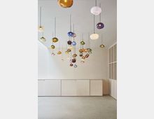 Minimalistischer Innenraum mit Hängelampen aus buntem Glas in unterschiedlichen Formen, die an langen schwarzen Kabeln von der Decke hängen. Der Raum hat helle Wände, eine einfache Holzverkleidung und einen Boden aus poliertem Beton.