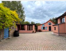 Ein gepflasterter Hof mit mehreren Gebäuden in warmen Rosa- und Backsteintönen. Links steht ein Gebäude mit einer blauen Holztür und gelb-grünem Efeubewuchs, rechts ein Wohnhaus mit blauen Fensterläden und rotem Ziegeldach.