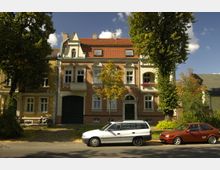 Fassade von historischen Wohnhäusern mit roten Ziegeldächern und verzierten Fenstern; vor den Gebäuden stehen zwei Autos und ein Baum wirft Schatten auf die Straße.