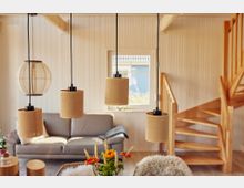 Gemütliches Wohnzimmer mit Holzverkleidung, einer grauen Couch, hängenden Lampen mit Stoffschirmen, einer Holztreppe im Hintergrund und Dekoration wie Blumen und einem Fellüberwurf. Ein Fenster mit Tageslicht sorgt für eine helle Atmosphäre.