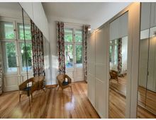 Ein heller Raum mit Holzfußboden, großen Fenstern mit floralen Vorhängen und einem Blick ins Grüne. Der Raum ist mit einem hohen, weißen Schrank mit Spiegeltüren und zwei Rattanstühlen ausgestattet.