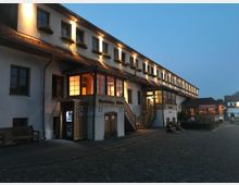 Großer, zweistöckiger Gebäudekomplex im traditionellen Baustil mit weißer Fassade, roten Dachziegeln und Blumenkästen vor den Fenstern. Die Beleuchtung und Holzverkleidungen betonen Eingänge und Fenster, während der gepflasterte Vorplatz von Abenddämmerung umgeben ist.