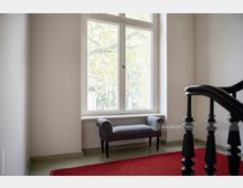 Heller Treppenhausbereich mit einem großen Fenster, vor dem eine graue gepolsterte Sitzbank steht; der Boden hat einen roten Teppich, und ein schwarzes Geländer ist teils sichtbar.