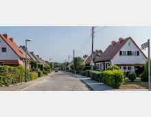 Wohnstraße mit gepflasterter Fahrbahn, gesäumt von Einfamilienhäusern mit weißen Fassaden und roten Satteldächern. Die Vorgärten sind gepflegt und durch grüne Hecken abgegrenzt, im Hintergrund sind Bäume und eine leichte Anhöhe zu sehen.