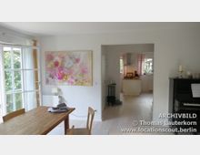 Wohnzimmer mit hellen Holzböden und weißen Wänden, dekoriert mit einem großen, bunten Blumenbild. Der Raum führt zu einer offenen, hellen Küche im Hintergrund; links gibt es eine große Fensterfront, rechts steht ein schwarzes Klavier.