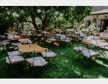 Ein Biergarten mit Holztischen und Klappstühlen, die mit Kissen ausgestattet sind, auf einer Wiese unter schattenspendenden Bäumen. Im Hintergrund ist ein Backsteingebäude mit Pflanzen und Dekorationen zu sehen.