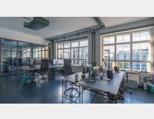 Ein modernes Großraumbüro mit mehreren Schreibtischen und Bürostühlen, großen Monitoren und Schreibtischlampen. Durch große Fenster mit Sprossen fällt Tageslicht ein und bietet einen Blick auf umliegende Gebäude; im Hintergrund sind gläserne Besprechungsräume sichtbar.