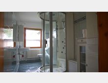 Badezimmer mit einer runden, verglasten Duschkabine in der Mitte, einem Waschbecken links, einem Fenster mit Holzrahmen im Hintergrund und einem weißen Heizungskörper darunter. Auf der rechten Seite befinden sich ein weißer Schrank und eine Wäschetruhe.