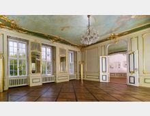 Prunkvoller Innenraum eines historischen Gebäudes mit goldverzierten Wänden, hohen Fenstern und einer bemalten Decke. Ein großer Kristallleuchter hängt von der Decke, und doppelflügelige Türen führen zu einem angrenzenden Raum.