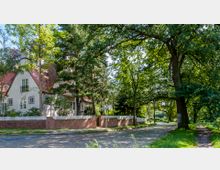 Einfamilienhaus mit weißer Fassade und rotem Satteldach, umgeben von einem Garten und einer Ziegelmauer mit weißem Zaun in einer baumreichen, ruhigen Wohngegend. Im Vordergrund eine gepflasterte Straße und ein schattiger Gehweg unter großen Bäumen.