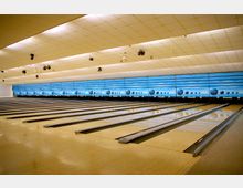 Innenraum einer modernen Bowlingbahn mit mehreren parallelen Bahnen und glänzendem Holzboden. Im Hintergrund befinden sich Wurfziele mit blauer, wellenförmiger Gestaltung und aufgedruckten Bowlingkugel-Motiven, während die Decke mit quadratischen Paneelen ausgestattet ist.