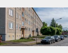 Mehrstöckiges Wohngebäude mit heller Fassade und mehreren Eingängen, entlang einer gepflasterten Straße mit parkenden Autos. Im Vordergrund sind Grünflächen, Bäume und Straßenlaternen erkennbar, im Hintergrund weitere Wohnhäuser und Bäume.