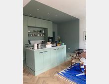 Eine moderne Wohnküche mit grün gestrichenen Einbauschränken und einer Kücheninsel in hellem Grün mit weißer Arbeitsplatte. Der Raum hat einen Fischgrät-Holzboden, eine minimalistische Einrichtung und einen blau-weiß gemusterten Teppich unter einem Esstisch mit Holzstühlen.