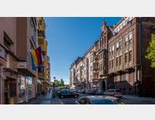 Straßenszene mit Wohn- und Geschäftsgebäuden, darunter ein modernes Gebäude mit Balkonen links und ein historisches Backsteingebäude mit dekorativen Fenstern rechts. Autos parken am Straßenrand, und Gehwege sind erkennbar, während blauer Himmel die Szene überlagert.