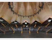 Großer moderner Veranstaltungsraum mit runder Sitzanordnung, ansteigenden Sitzreihen und einer auffälligen, geometrischen Holzdecke mit integrierter Beleuchtung. Über der Mitte des Raums hängt ein kreisförmiges Beleuchtungssystem, und die Wände sind in dunklen Tönen gehalten.