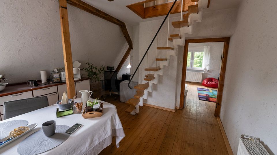 Offener Wohn- und Essbereich mit Holzdielenboden, weiß verputzten Wänden und sichtbaren Holzbalken; links eine Küchenzeile und ein Esstisch. In der Mitte führt eine schmale Holztreppe zu einer Dachluke, rechts ein Durchgang in ein Zimmer mit Fenster und kleinem Teppich.