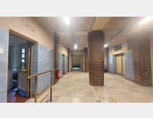 Ein Flur in einem historischen Gebäude mit Wänden aus roten Ziegelsteinen und blau gefliesten Bereichen. Der Boden ist mit hellen Fliesen bedeckt, und an der Decke sind schlichte Leuchtstofflampen angebracht; ein Schild mit der Aufschrift „Keine Schwimmhalle“ ist im Hintergrund sichtbar.