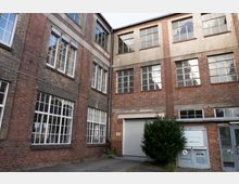 Innenhof eines älteren Industrie- oder Fabrikgebäudes aus rotem Backstein mit großen, vielfach unterteilten Fensterfronten und verputzten Wandbereichen. Unten sind ein Rolltor, eine Seitentür und ein kleiner Strauch vor der Fassade zu sehen.