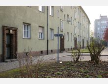 Mehrstöckige Wohnhäuser mit grün verputzter Fassade, flankiert von Klinkerziegeln um die Eingangsbereiche; davor ein Gehweg, einige kahlen Sträucher und ein mit Laub bedecktes Beet. Im Hintergrund sind weitere Wohngebäude erkennbar.