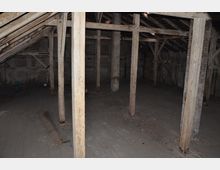 Dunkler Dachboden mit sichtbaren Holzbalken, schrägem Dach und einem unlackierten Holzboden. Die Wände und die Konstruktion bestehen aus Holz und zeigen Spuren von Verwitterung.